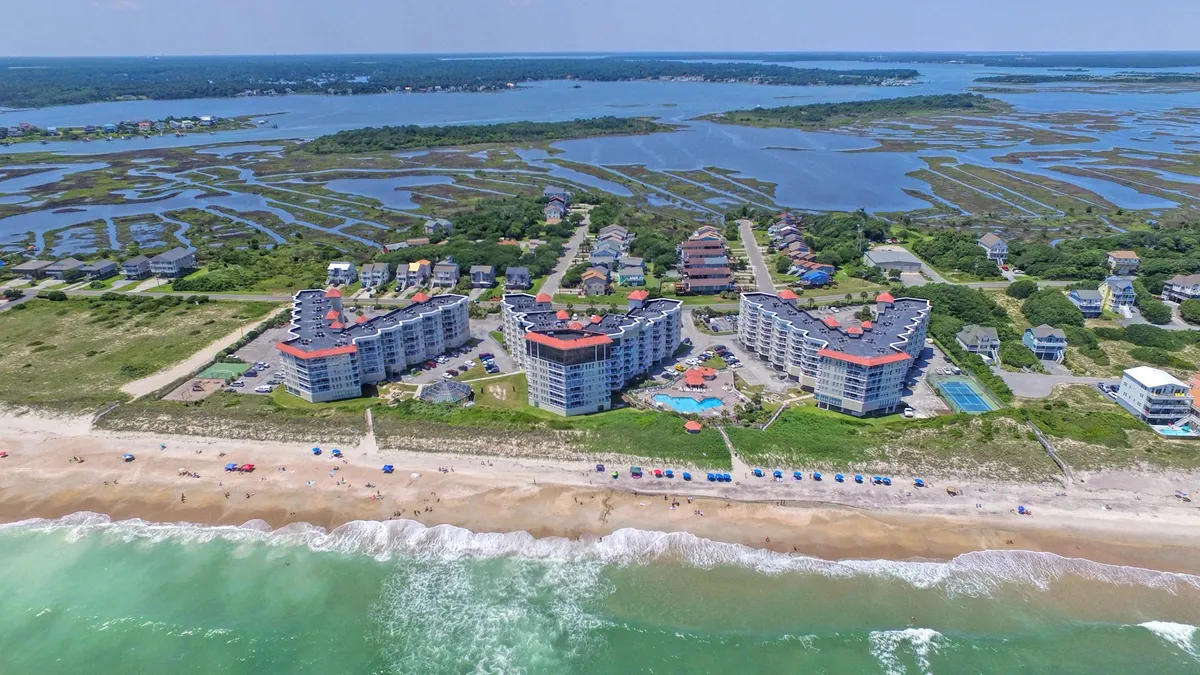 A vibrant beachfront scene featuring oceanfront condos, a sparkling swimming pool, colorful beach umbrellas, and marshlands in the distance. The image captures guests swimming, sunbathing, beachcombing, and enjoying both the pool and the beach. The dynamic setting hints at the easy transition from condo comfort to outdoor fun.