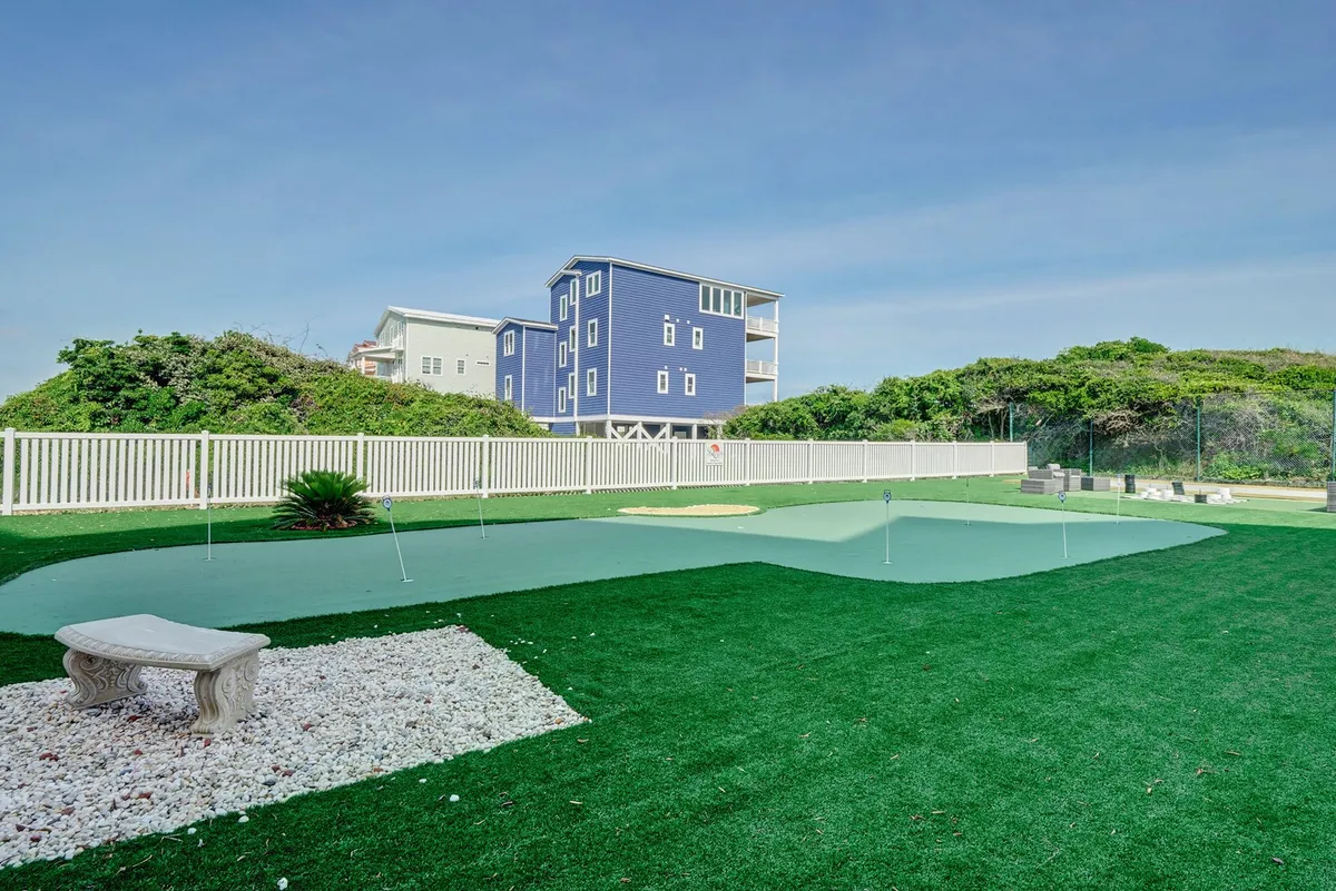 A neatly maintained green artificial turf hosts a golf putting area, with a stone bench and surrounding trees and a blue house in the background. The setting is playful, casual, and perfect for socializing or relaxing between meals.