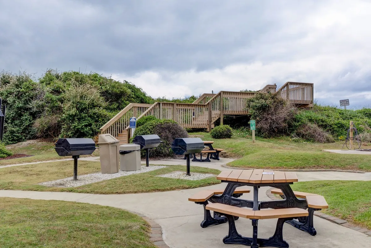 The scene shows picnic tables and barbecue grills set among greenery, with stairs leading up to a deck. The area is designed for outdoor barbecuing, communal meals, and social gatherings, ideal for both families and groups of friends.