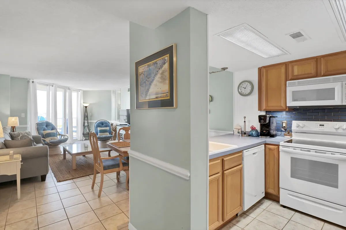 This image captures an open-concept living space that cohesively unites the kitchen area, a dining table, and a living area with a welcoming sofa. The room is filled with natural light, accentuating the modern decor and comfortable furnishings. Subtle coastal touches and hardwood floors set a relaxed, breezy tone, while the arrangement is clearly designed for both social gatherings and relaxation.