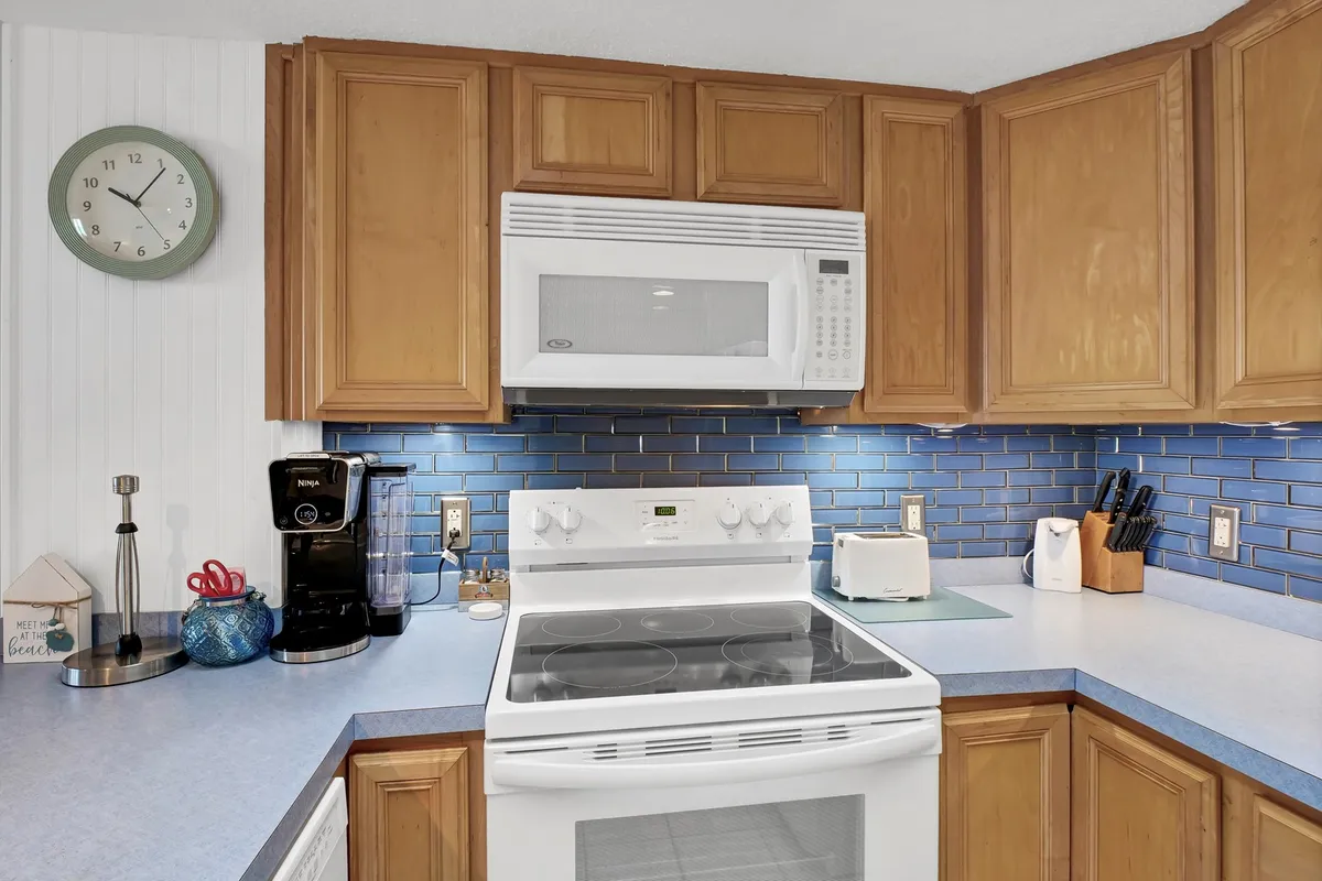 A closer view of the kitchen area reveals a white stove, blue ocean-inspired backsplash, coffee maker, and wooden cabinets. The intimate scale highlights the kitchen's style and functionality—ideal for prepping snacks or sunrise coffee against a fresh, coastal palette.