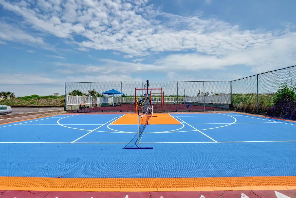 This vibrant exterior image captures a multi-use court area with a basketball hoop, tennis net, and adjacent playground equipment. The court is brightly colored and spacious, set against a backdrop of greenery, inviting guests to engage in a variety of sports and recreational activities.