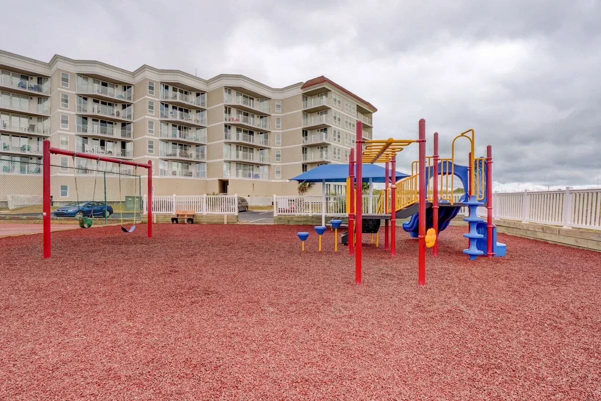 This exterior image spotlights a cheerful playground area, complete with colorful playground equipment, multiple swings, and a safe rubber ground covering. The space is surrounded by greenery, providing a welcoming and secure spot for families and children to enjoy outdoor play.
