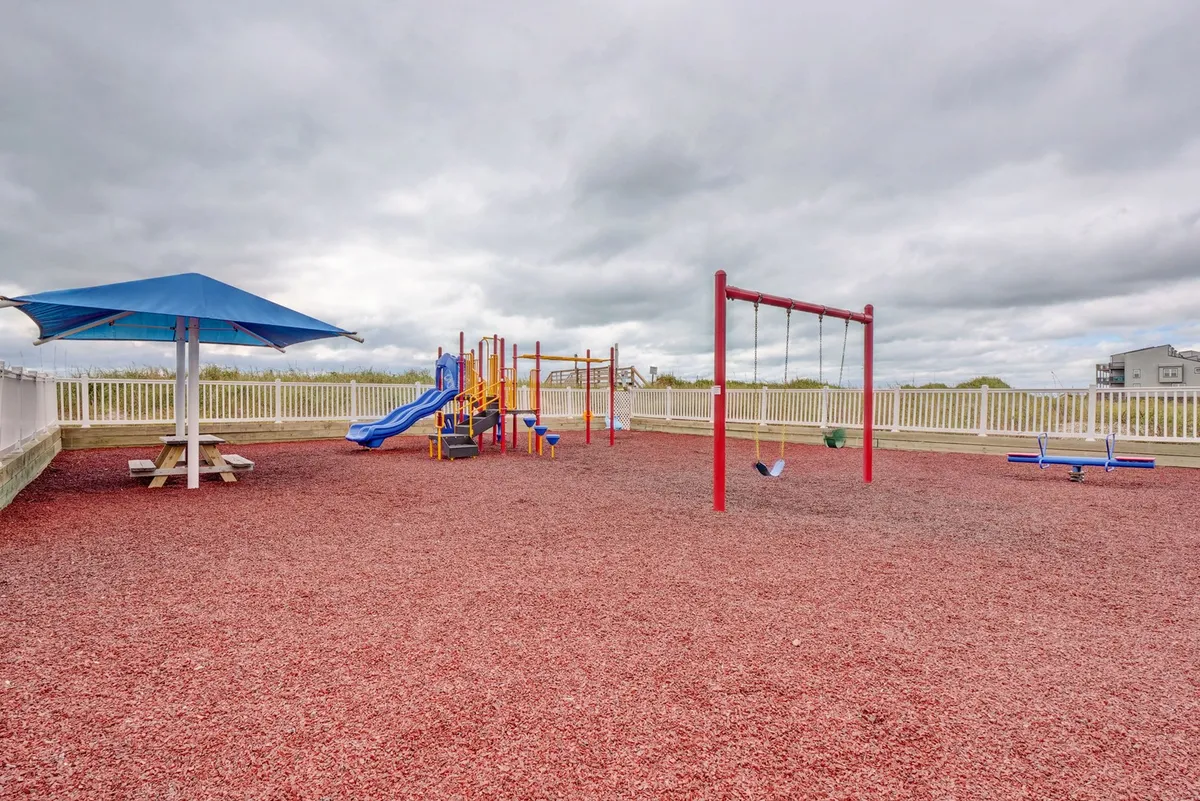A vibrant playground with colorful equipment and a swing set sits beside a shaded picnic area. Families and children are seen playing and interacting, creating a joyful, community-oriented environment.
