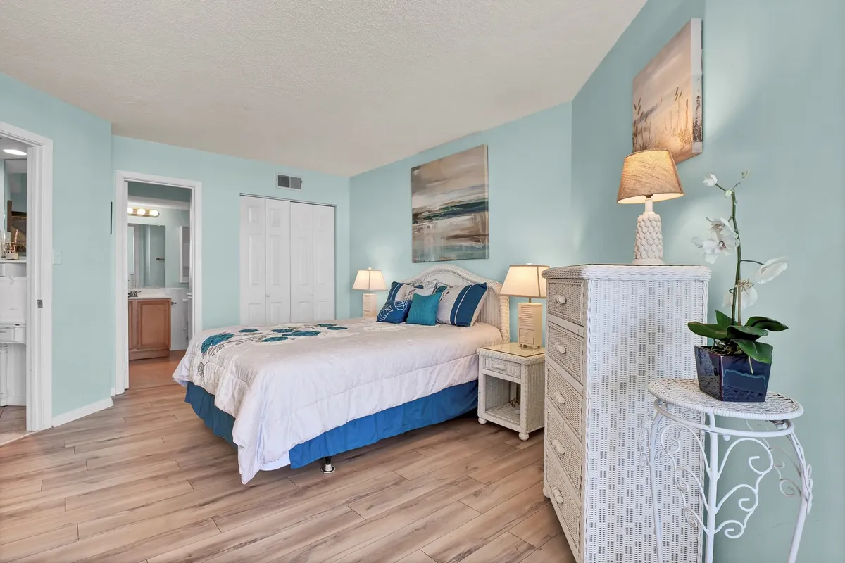 A tranquil bedroom with a cozy bed adorned with decorative pillows, artwork on the wall, and a nightstand with a lamp. Wicker furniture adds a homey, inviting feel, and natural light streams in to create a serene atmosphere.