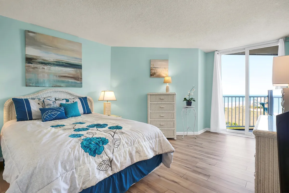 This image highlights a tranquil bedroom featuring a bed with a floral design, a large window offering an ocean view, and a nightstand with a lamp. The space is filled with natural light and exudes a peaceful, restful atmosphere—ideal for relaxing after a day by the shore.