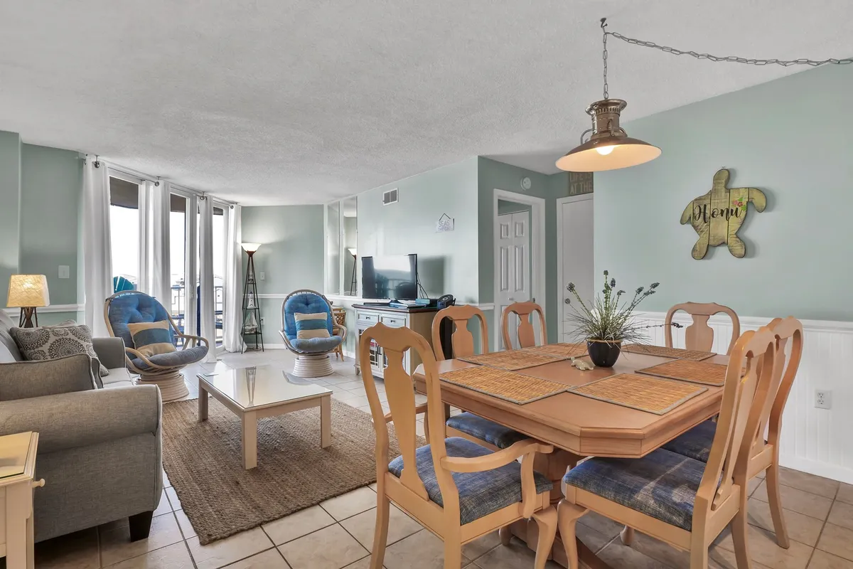 A spacious, open-plan living and dining area featuring a large dining table surrounded by chairs, a comfy sofa, lounge chairs, a television, and decorative wall art. The space is filled with natural light and designed to encourage relaxation and socialization.