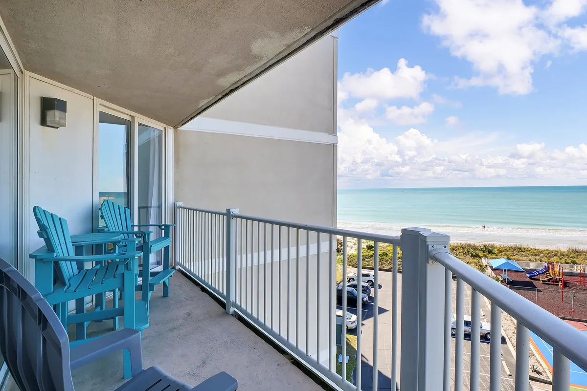 This image features a balcony with comfortable seating overlooking the ocean and beach. The tranquil outdoor space is perfect for enjoying a meal or drinks while taking in sweeping views of the water and sky. The ambiance suggests relaxation and socialization, ideal for dining or unwinding.