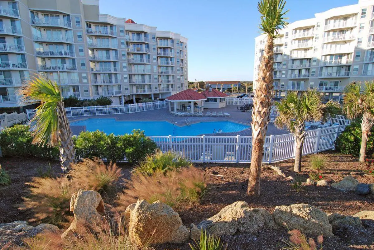 A sparkling swimming pool surrounded by palm trees, with lounge chairs and a poolside gazebo. Modern apartment buildings can be seen in the background, creating a resort-like atmosphere. The area is designed for relaxation and socializing, with guests enjoying the pool and outdoor spaces.