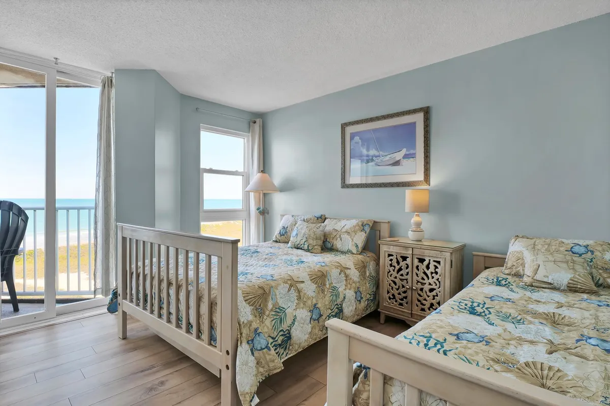 A bright guest bedroom with two beds adorned in floral bedding, a decorative lamp, artwork on the wall, and a window framing an ocean view. The room feels fresh and versatile, suitable for both families and small groups, and encourages togetherness and relaxation.