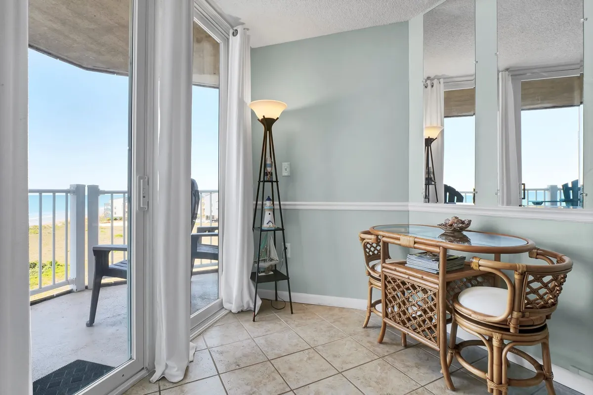 A cozy rattan dining set is positioned near a large window with an ocean view. A floor lamp and airy décor contribute to the relaxed, bright atmosphere, ideal for morning coffee, reading, or working with a tranquil backdrop.